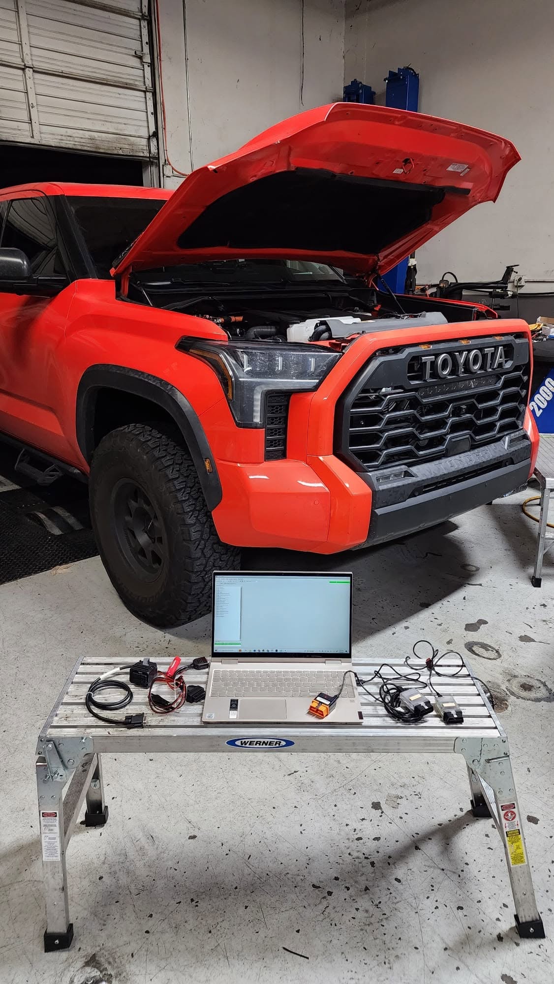 Orange Toyota Tundra with its hood open and a laptop, cables and tuning interfaces on a workbench in front.